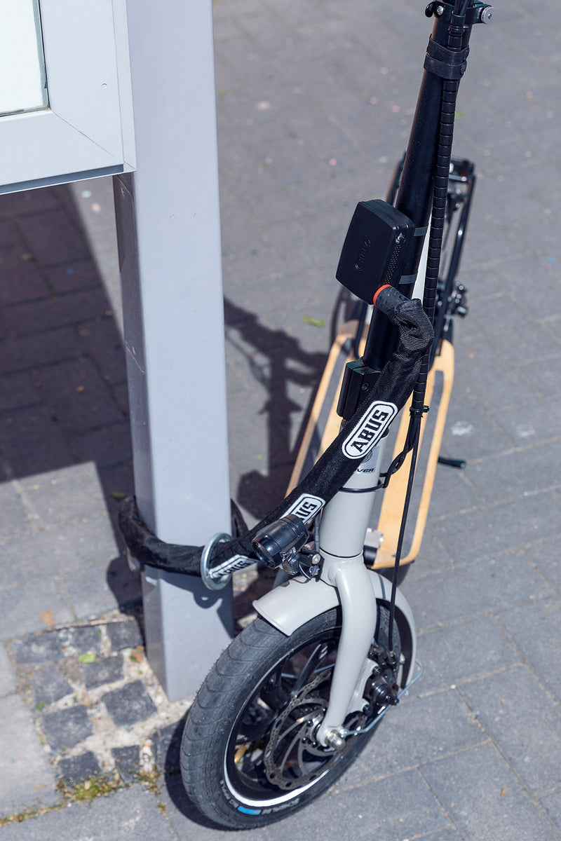 Load image into Gallery viewer, A folding electric scooter with a black frame and a light beige deck, parked next to a white post on a paved outdoor street and locked with loop chain and AlarmBox
