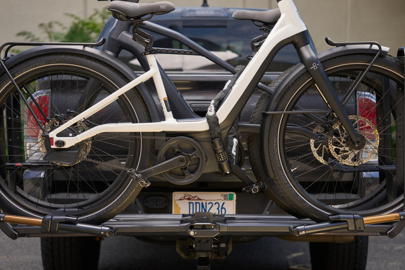 Load image into Gallery viewer, A white and black electric bicycle with rear racks, shown mounted on a hitch-style bike carrier attached to the back of a dark pickup truck. #color_black

