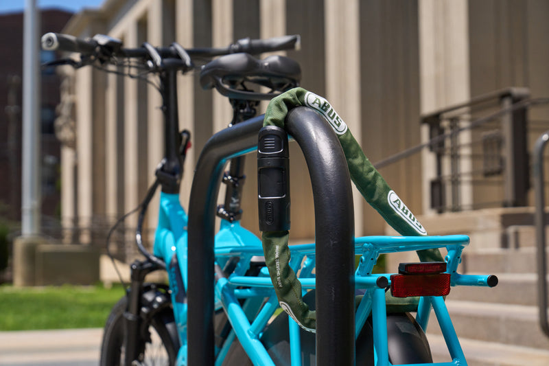 Load image into Gallery viewer, A turquoise electric cargo bike with a green ABUS lock wrapped around its black front rack, parked outdoors in front of a beige building. #color_jade green
