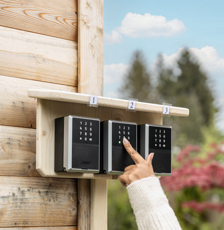 An ABUS Keygarage on a wooden wall with a hand pressing the keypad, surrounded by greenery.